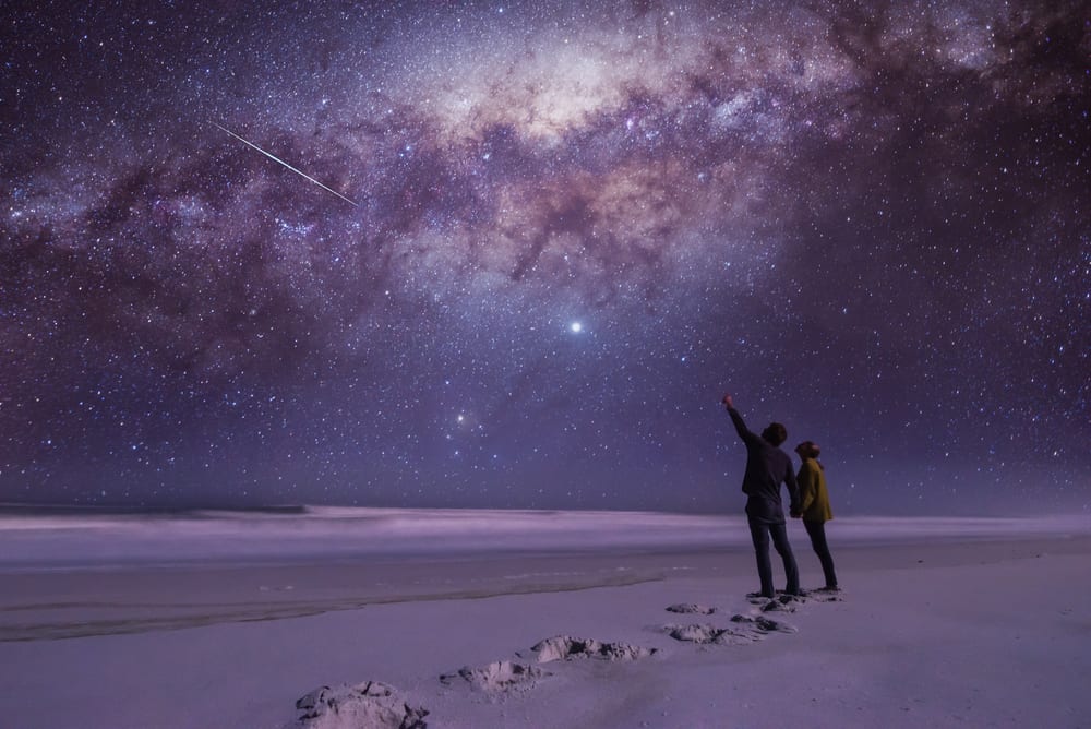 couple stargazing in a beautiful open spot with a blue and purple milkway above them